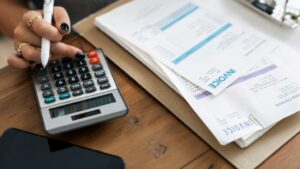 Person using a calculator next to a stack of invoices and financial documents, illustrating the process of reconciling accounts.