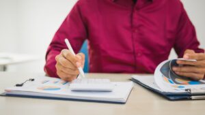 A person in a maroon shirt working at a desk with financial documents, using a calculator and pen—representing tasks related to payroll compliance.