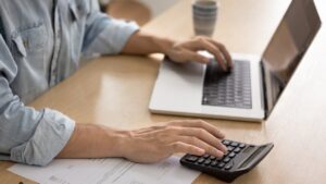 Small business owner using a laptop and calculator to manage accounting and bookkeeping tasks, with financial documents on desk.