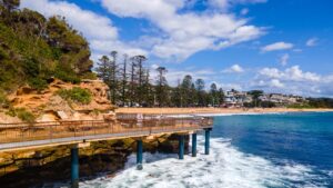 Elevated coastal walkway over ocean waves at Terrigal on the Central Coast, New South Wales, with sandstone cliffs, pine trees, a sandy beach, and residential buildings under a partly cloudy sky.