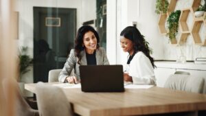 Two professional women collaborate on accounts payable management tasks while working together at a laptop in a modern office setting, reviewing financial documents and discussing payment processes.