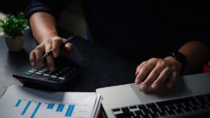 Small business owner managing payroll for small business, calculating figures on a calculator while reviewing financial reports and working on a laptop.